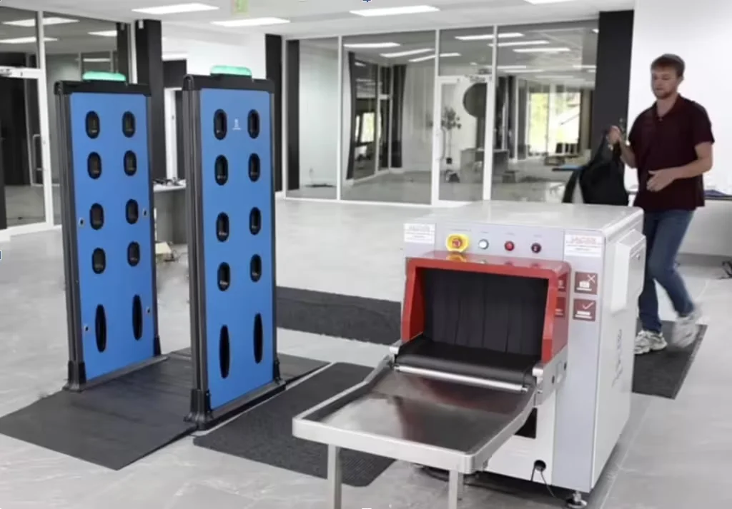 a man wearing black t-shirt and holding a bag about to enter Athena's WDS and X-ray baggage scanner for weapons screening
